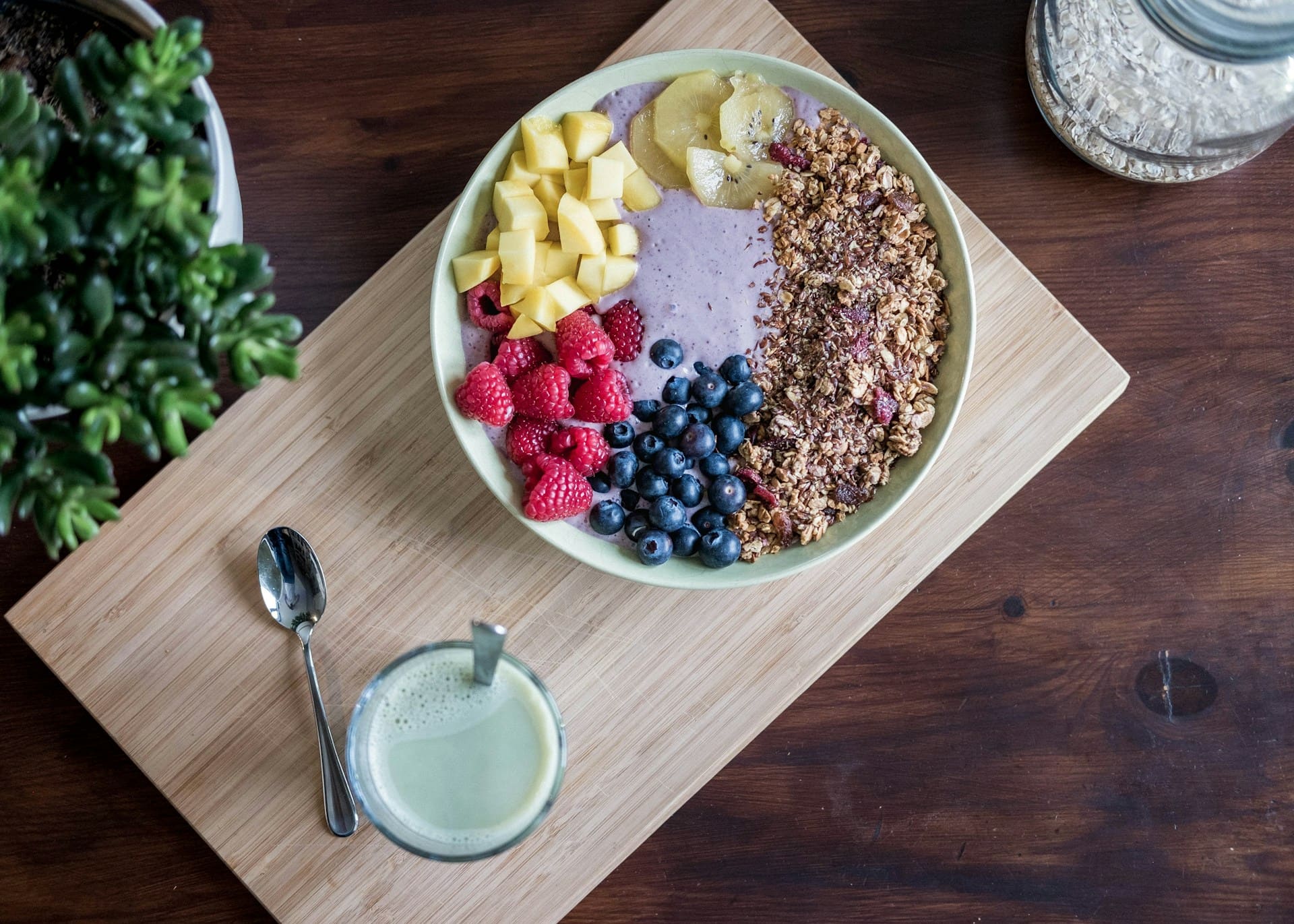 Un bol cu fructe proaspete și granola pe o masă de lemn, alături de un pahar cu smoothie verde și o linguriță.